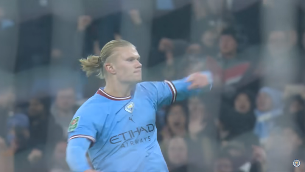 Erling Haaland in blue t-shirt gesturing with his hands, the audience behind him